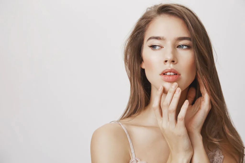 Studio shot of affective romantic caucasian woman with clean skin gently touching chin and looking aside with dreamy expression, standing over gray background.